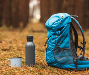 A blue camping backpack sits on a dry forest floor with a water bottle and enamel mug on the ground beside it.